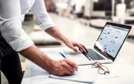 Project manager standing in modern industrial factory, working on laptop.