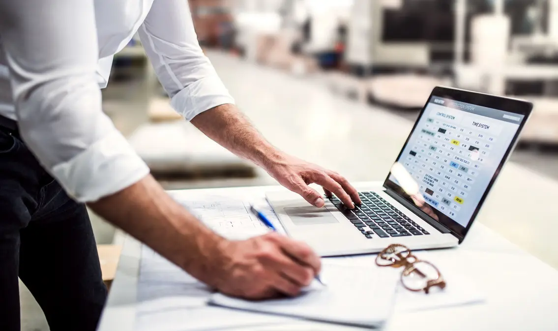 Project manager standing in modern industrial factory, working on laptop.